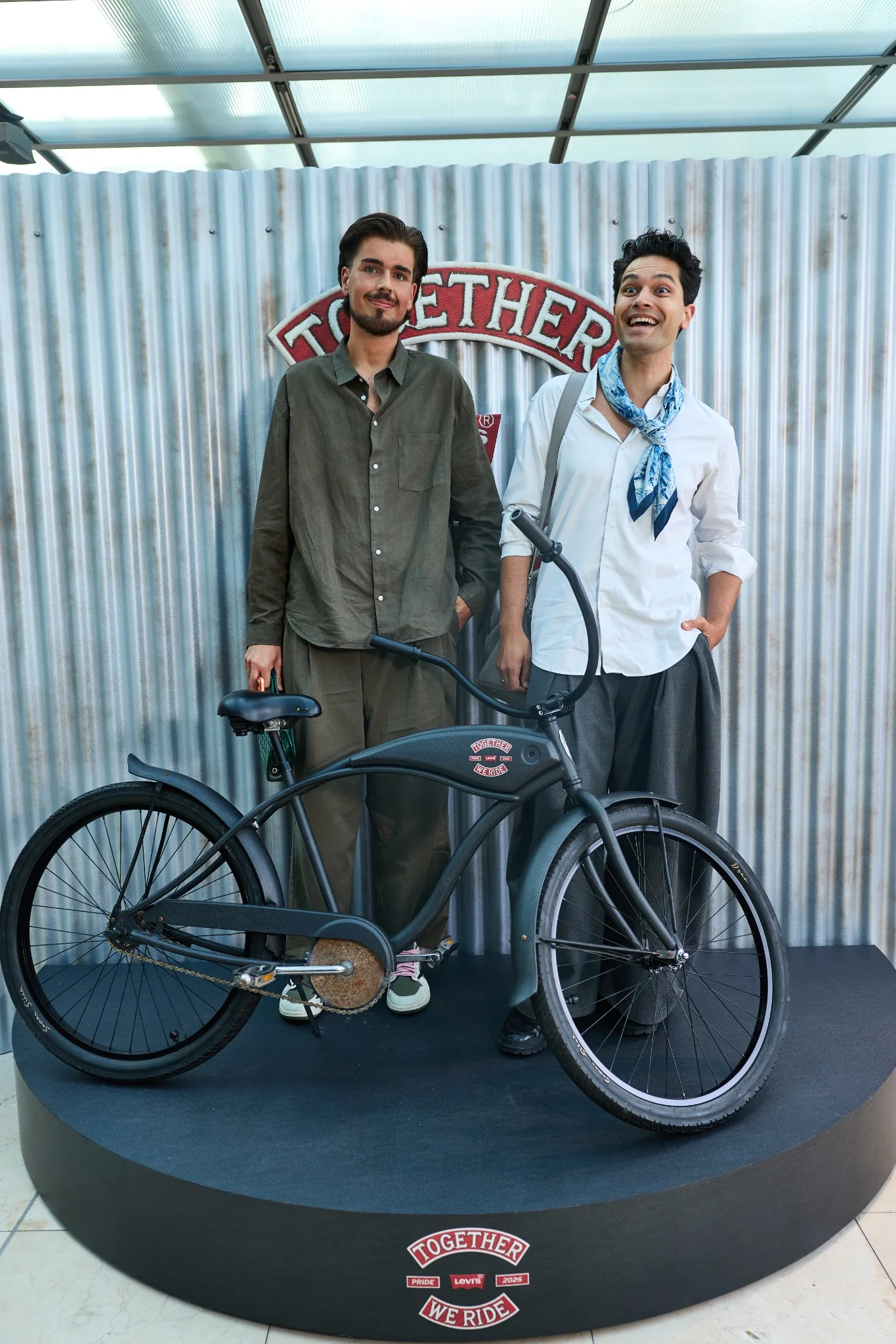 Two guests side by side behind the bike in front of the corrugated wall