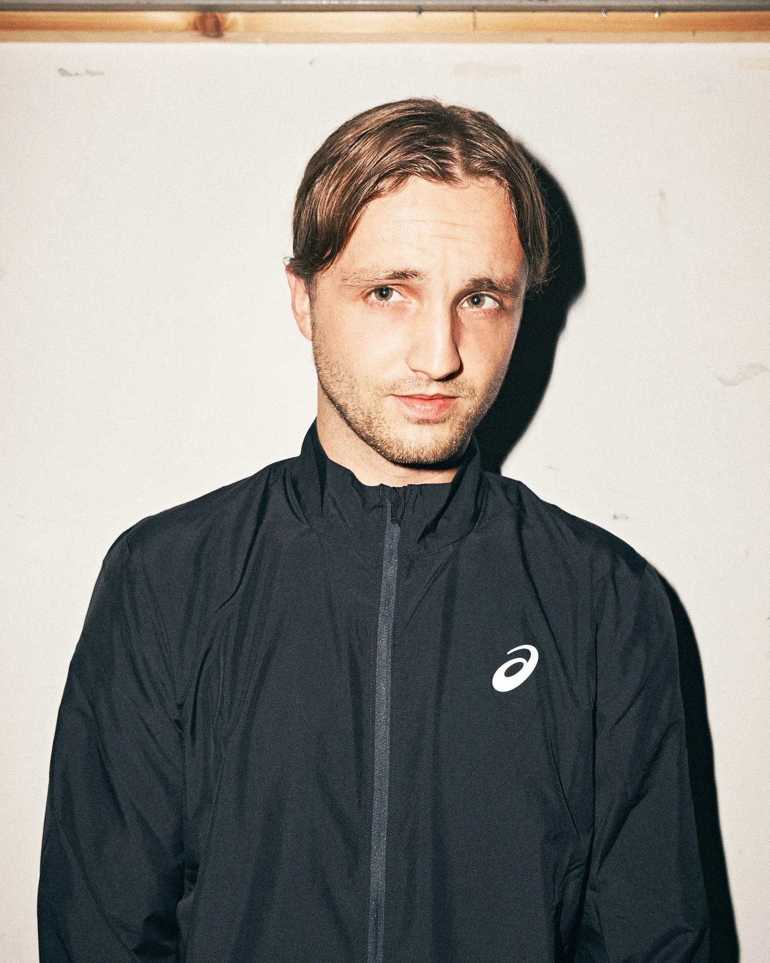 Portrait of Kevin in a black track jacket against a white wall