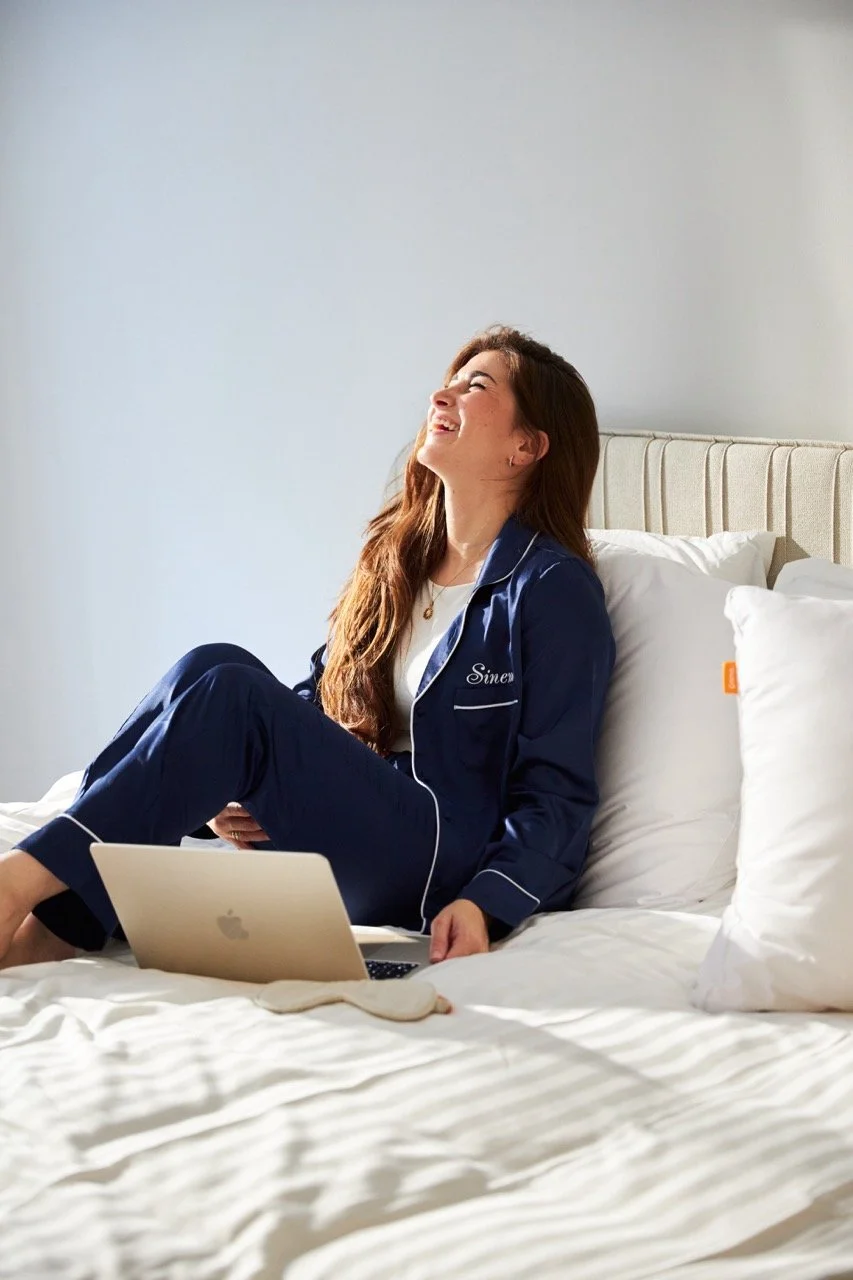 Woman in navy Emma sleepwear laughing on a bed with a laptop