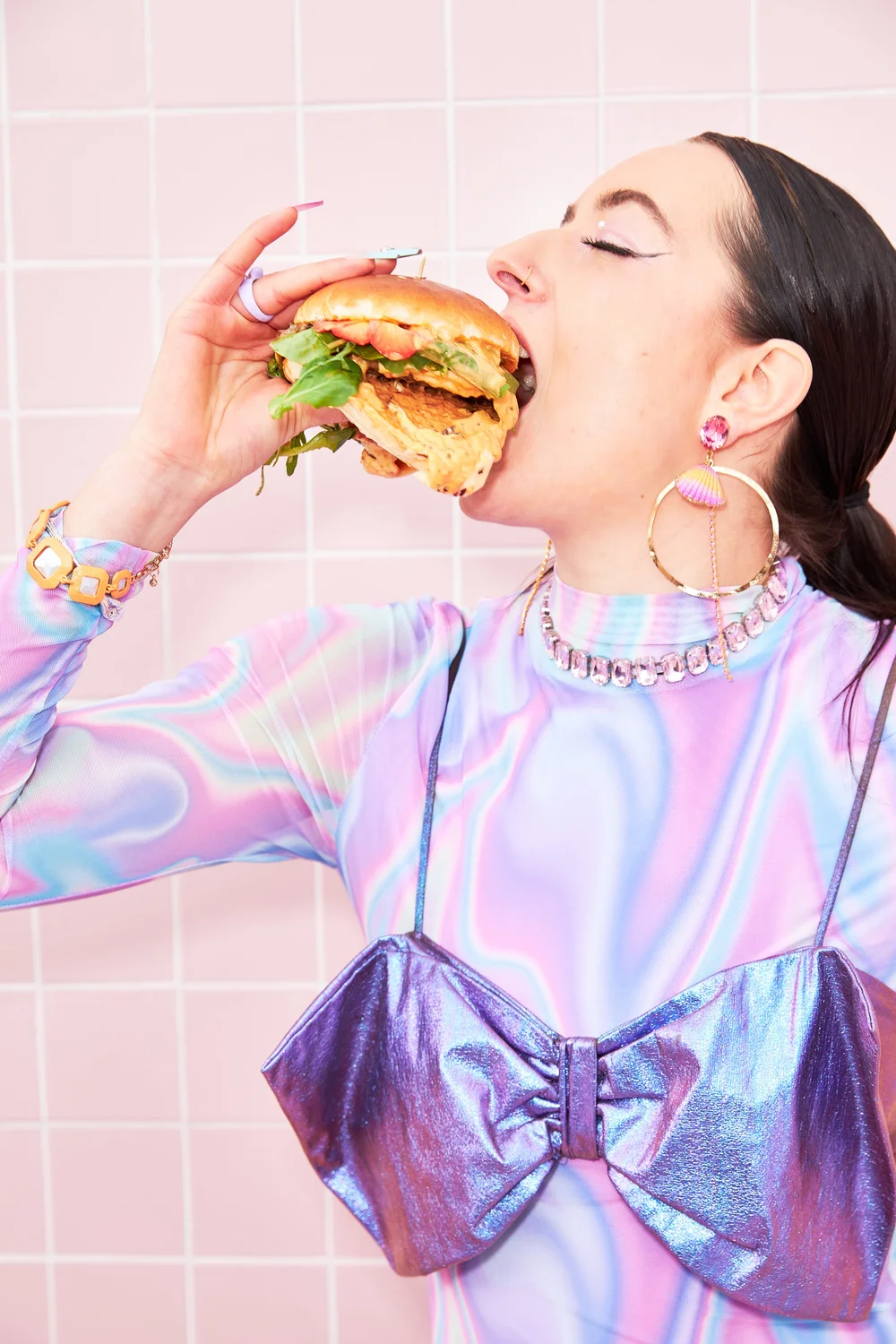 Woman in iridescent dress biting a burger against pink tile