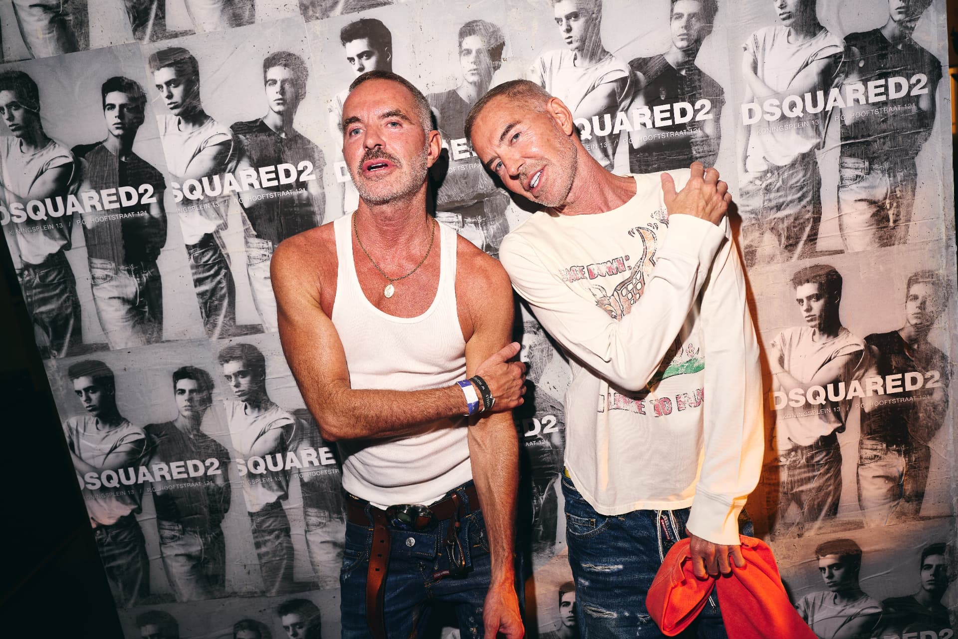 Dean and Dan Caten posing together in front of the DSQUARED2 poster wall
