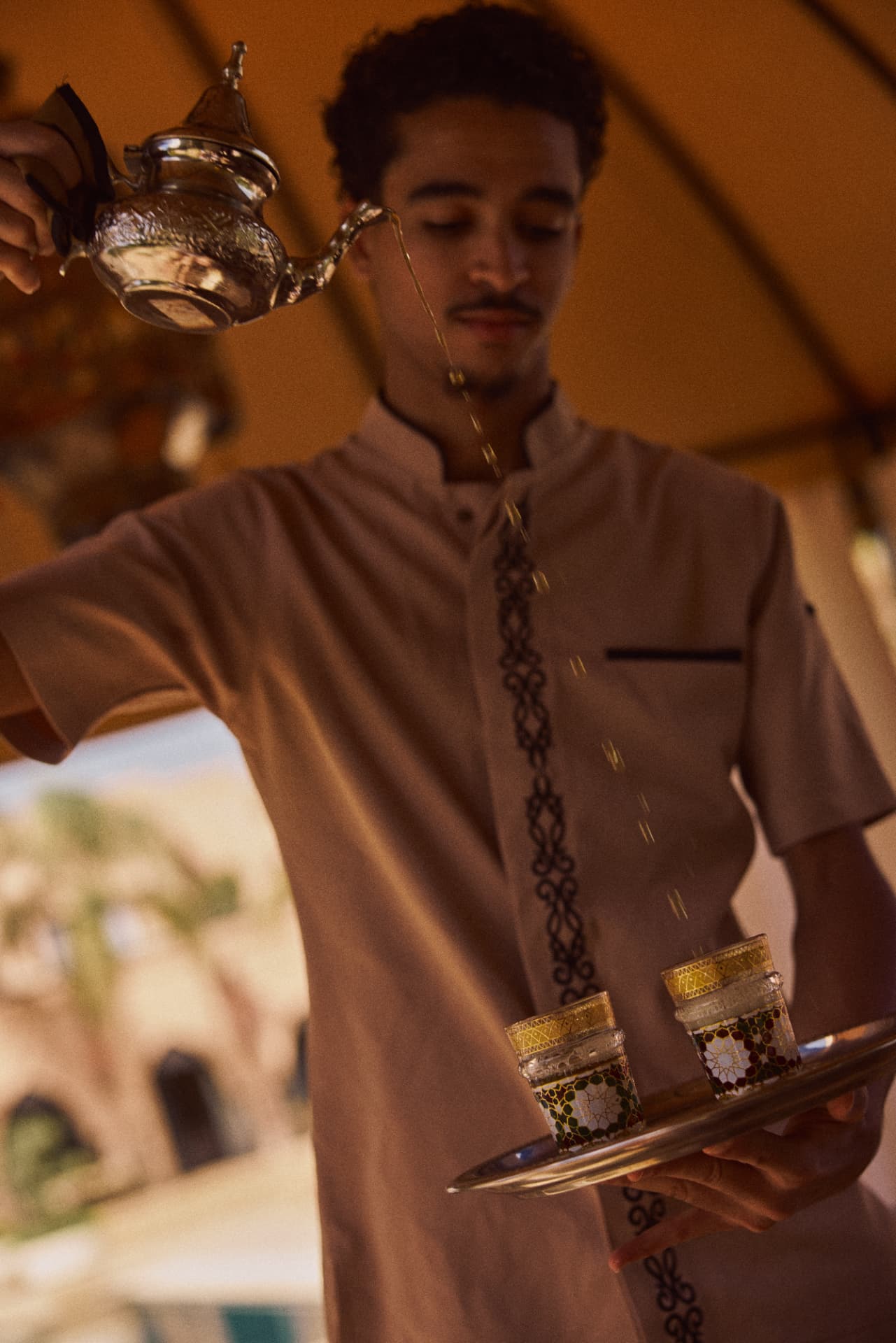 Waiter pouring mint tea from a silver pot onto a tray of glasses